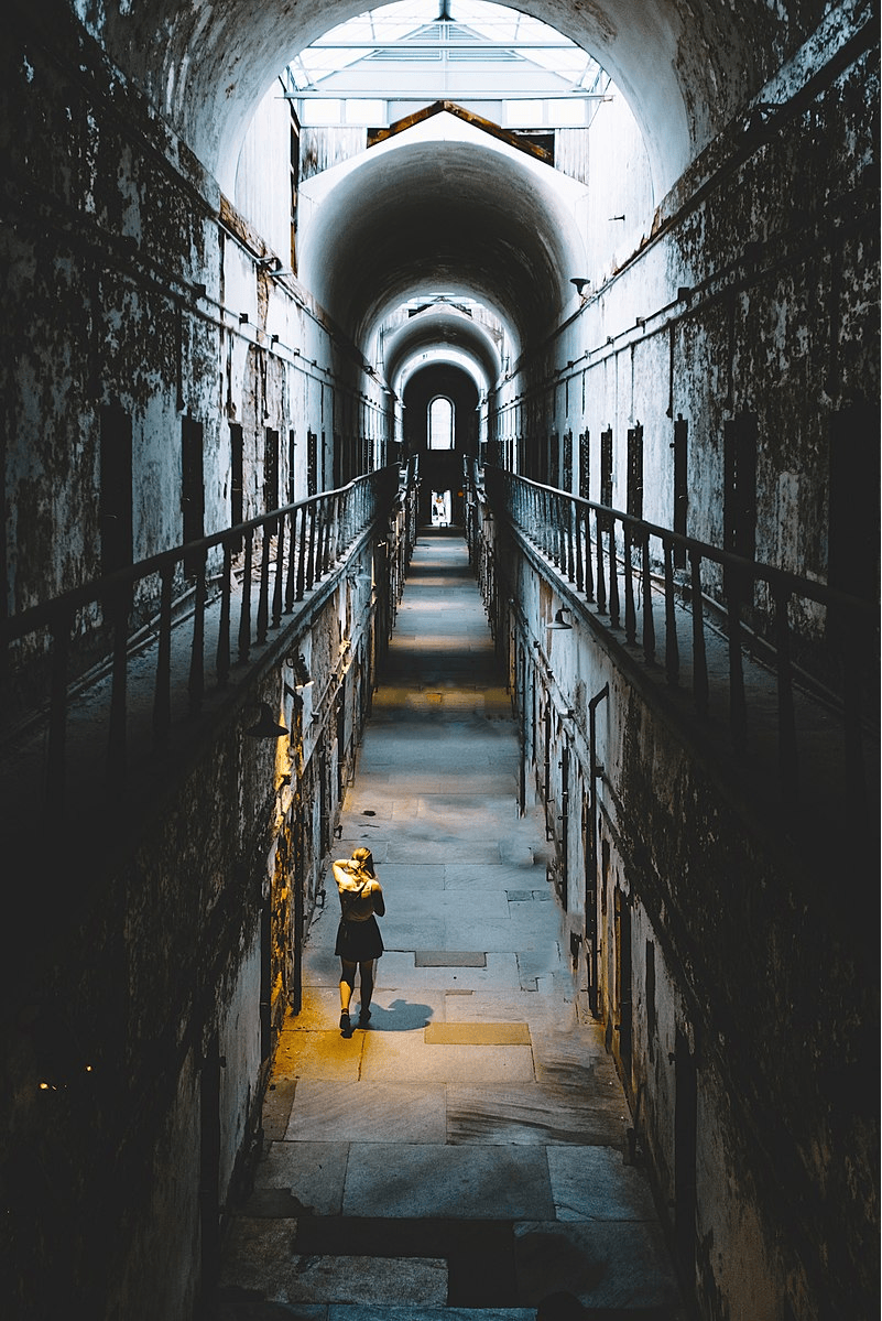 Person walking in a dim, narrow, arched corridor with light streaming from above.